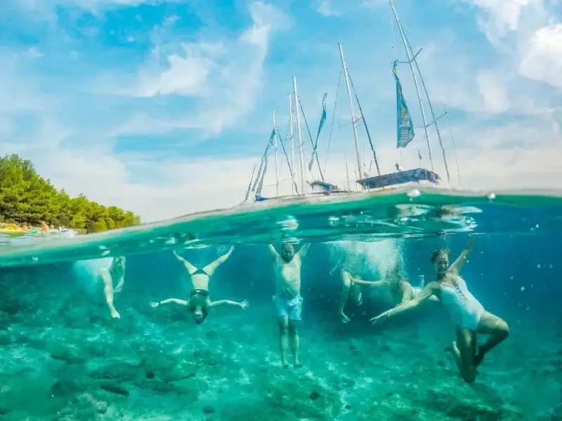 Split-level photo of MedSailors guests swimming underwater beneath anchored sailing yachts in clear Mediterranean water near a pine-lined shore.