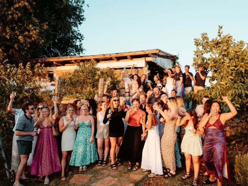 Large group of young travellers celebrating with drinks at a sunset dinner during a Greek island hopping trip from Athens. Social summer travel experience in Greece with friends enjoying local food, wine, and golden hour views on an affordable island sailing holiday.