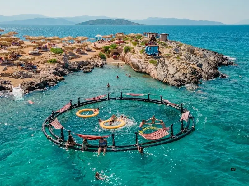 Aerial view of the Aponisos Beach blue lagoon on Agistri Island, Greece, featuring travelers swimming in bright turquoise water around a circular floating dock with hammocks, surrounded by rocky coastlines and beach clubs.