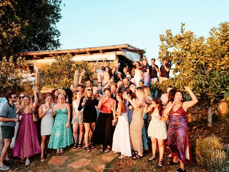 Group of MedSailors travellers celebrating together at a sunset island party in Croatia, raising glasses and enjoying a social crew night surrounded by trees and rustic decor on a Hvar Island vinyard— capturing the lively atmosphere of a MedSailors sailing holiday.