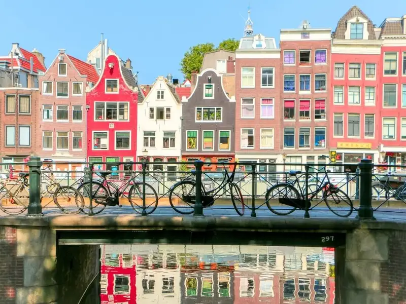 Colourful traditional canal houses line an Amsterdam canal on a bright day, with bicycles locked along a bridge railing in the foreground and reflections of the buildings shimmering in the water below. The scene captures Amsterdam’s walkable, bike-friendly vibe and classic canalside architecture, ideal for first-time Europe travellers looking for an easy city break with neighbourhood cafés, museums, and scenic strolls.