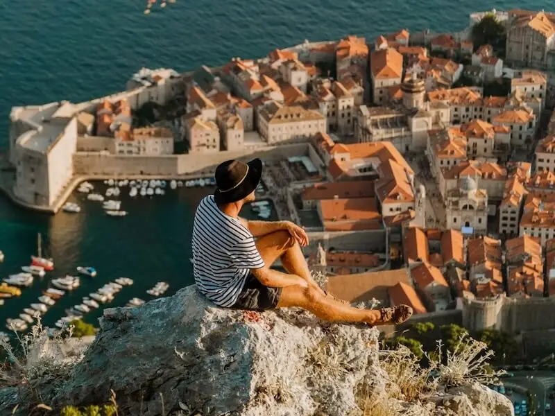 Traveller sitting on a cliff overlooking Dubrovnik Old Town and the Adriatic Sea on a clear day in Croatia.