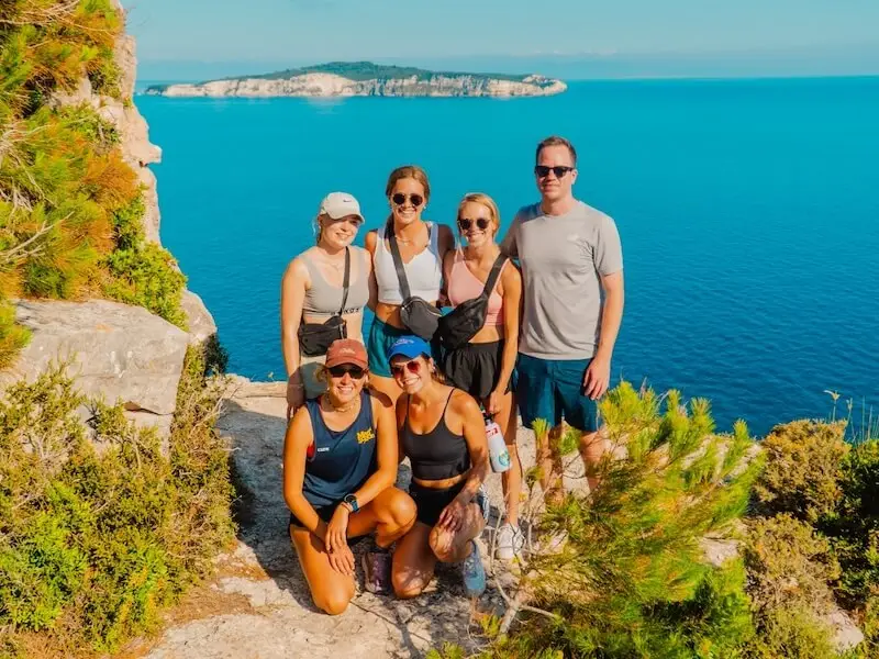 Group of MedSailors travellers posing on a cliffside hike near Tripitos Arch in Paxos, Greece, with clear blue sea and rugged coastline in the background.