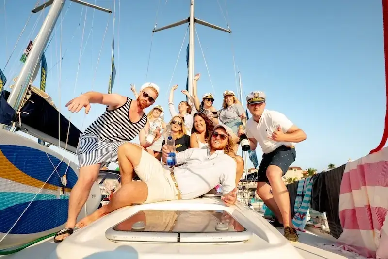 Young travellers on a MedSailors yacht celebrating during a sailing holiday, posing on deck in the sun with captain hats and drinks, anchored along a Croatia or Greece island-hopping route.