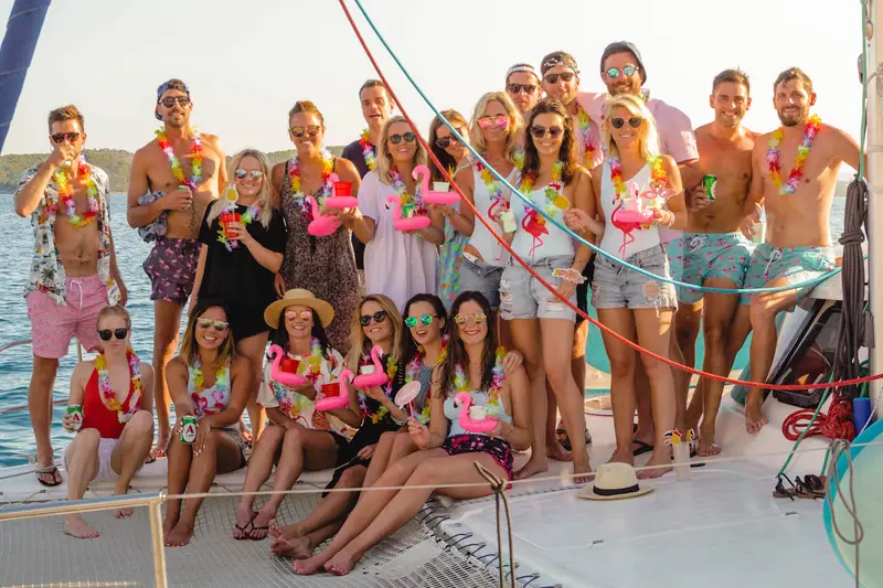 A group of travellers in their 20s or 30s sitting on a catamaran in the Mediterranean on a group island hopping tour. They are all wearing matching colouful outfits at sunset.