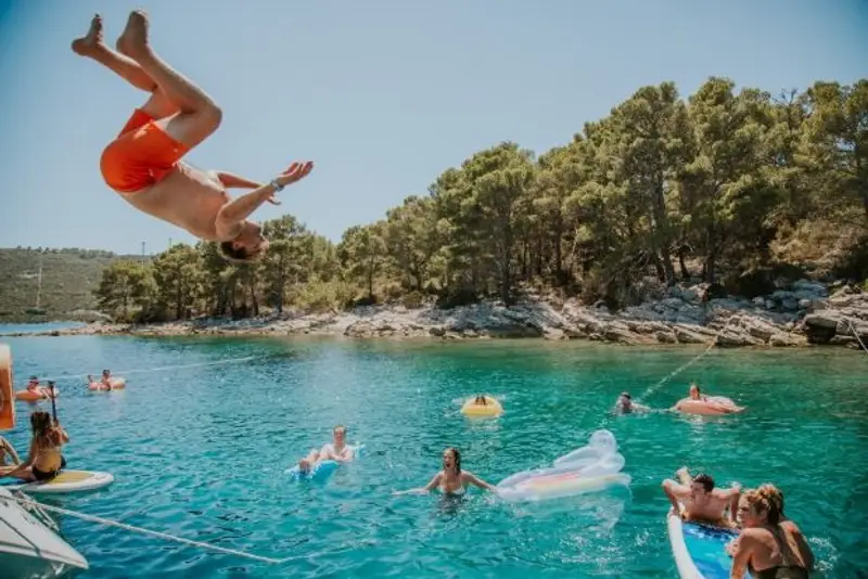 A daring traveller backflips off of a sailing yacht on an island hopping trip in the Mediterranean as other travellers look on.