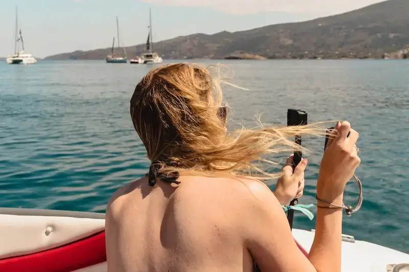 Woman with windswept hair capturing sailing footage on a GoPro while cruising on a MedSailors yacht in Greece, with anchored boats and hilly coastline in the background.