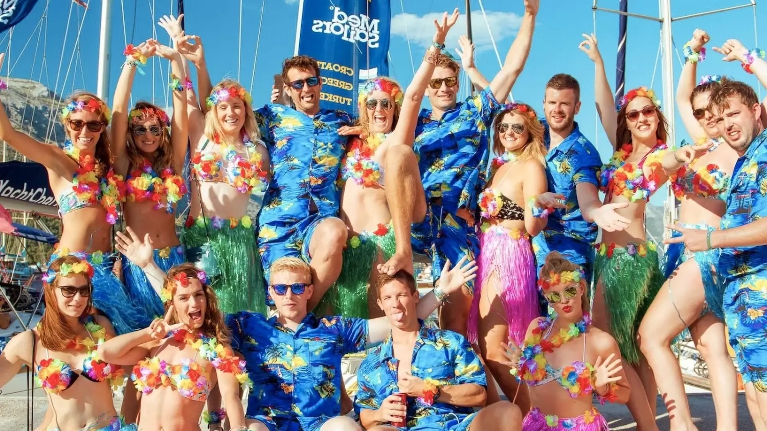Group of young adults dressed in tropical Hawaiian-themed outfits posing together on a docked yacht during a MedSailors sailing trip, celebrating in colourful leis, grass skirts, and matching shirts with festive energy.