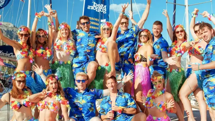 Group of young adults dressed in tropical Hawaiian-themed outfits posing together on a docked yacht during a MedSailors sailing trip, celebrating in colourful leis, grass skirts, and matching shirts with festive energy.