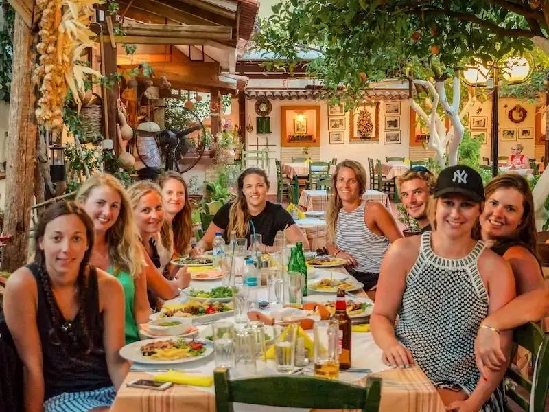 Group of female solo travellers and budget travellers exploring the Greek islands with MedSailors. Sitting at a table filled with Greek dishes in a colourful taverna.
