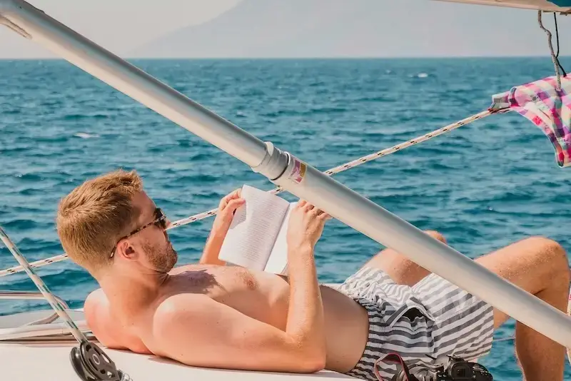 Man relaxing shirtless on a yacht deck reading a book during a MedSailors sailing holiday in Croatia, with calm blue seas and sunshine in the background.