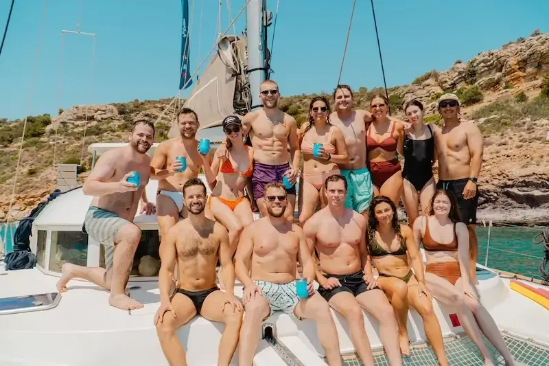 Group of guests posing on the deck of a catamaran in swimwear with drinks in hand, anchored in a secluded bay during a Greece yacht week with MedSailors.