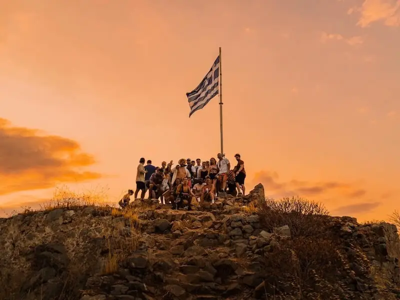 Group of MedSailors travellers gathered at the Greek flag viewpoint on Hydra Island during sunset, overlooking the Aegean Sea from a rocky hilltop.