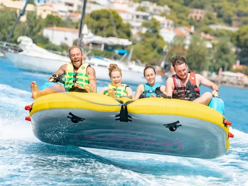 MedSailors guests riding a watersports on the sea in Greece, laughing and holding on during a high-speed boat tow with yachts in the background.