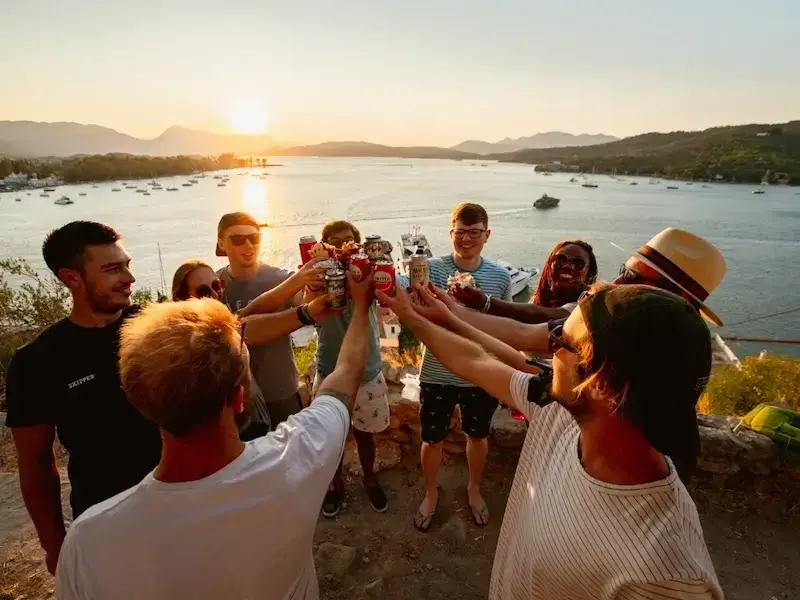 Group of budget backpackers cheers beers atop a viewpoint at sunset in Greece while on a Greek island hopping sail week with MedSailors.