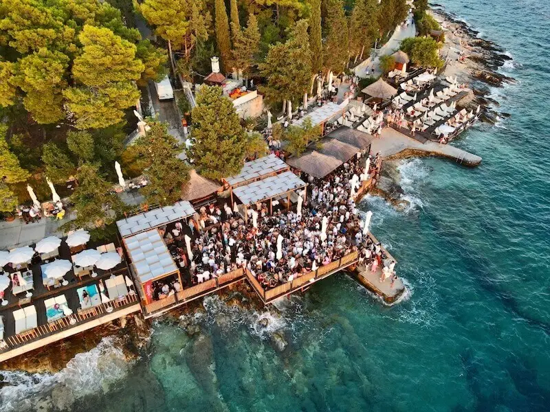 Aerial view of Hula Hula Bar in Hvar, Croatia, packed with MedSailors guests and partygoers along the waterfront, surrounded by turquoise sea and pine trees.