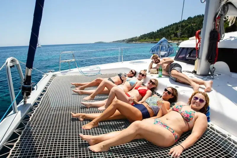 Group of young travellers sunbathing on the net of a MedSailors catamaran during a Croatia sailing holiday, surrounded by clear blue water and coastal views.