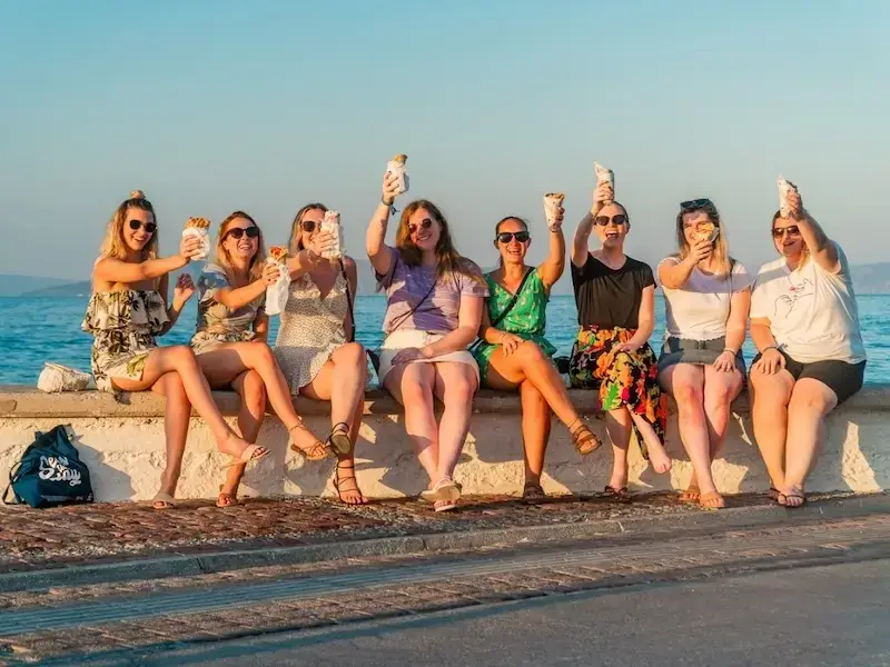 Group of female travellers enjoying a budget-friendly Greek meal of gyros by the seaside with an idyllic backdrop while on an adventure travel experience with MedSailors.