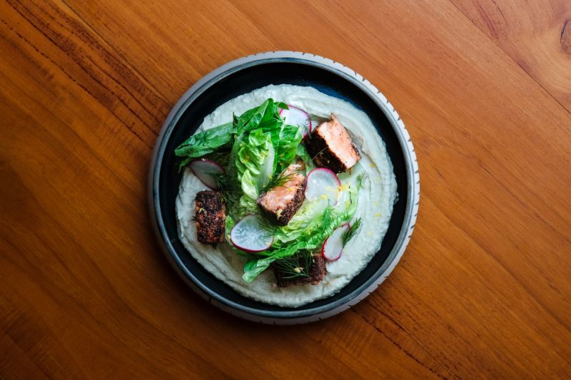 Spiced salmon pieces with salad leaves, sliced radish and creamy dressing in a ceramic bowl.