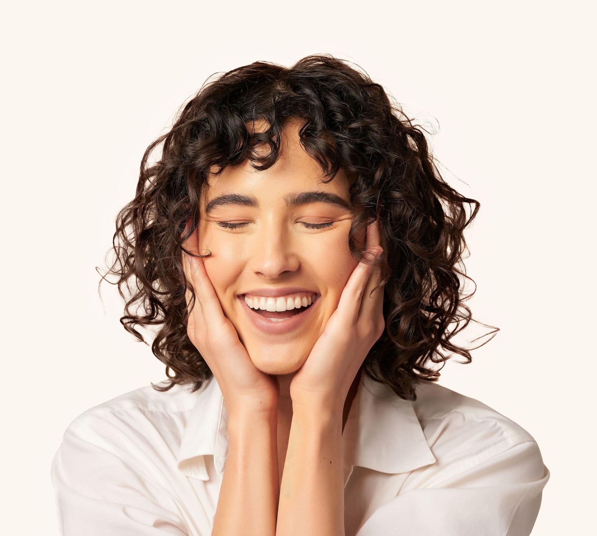 Photo of female with brunette curly hair smiling with eyes closed and hands on face