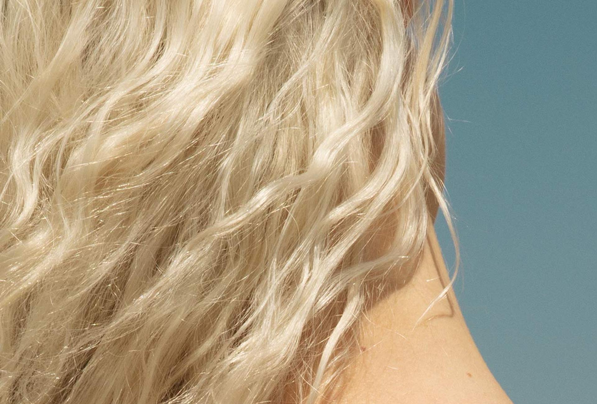 Image of the back of a model's head and neck showcasing blonde windswept frizzy hair with blue sky background