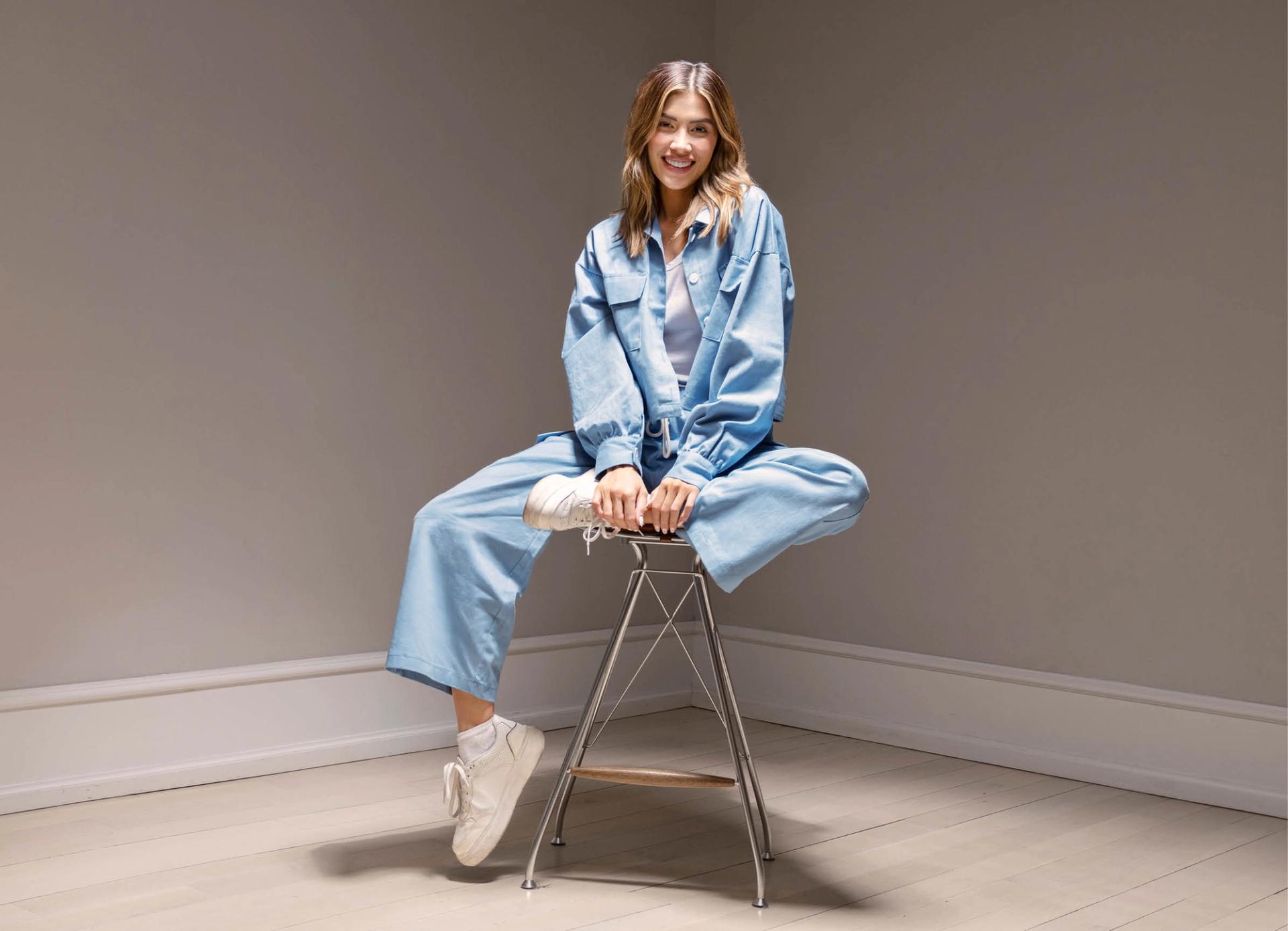 Michelle Salas in Blue Denim sat cross legged on stool smiling in corner of room