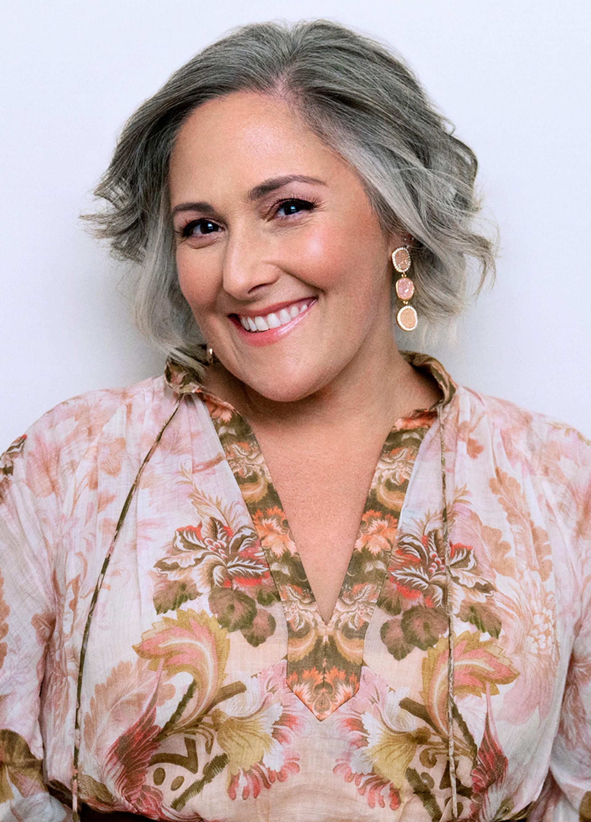 Ricki lake smiling directly at camera wearing a pink floral blouse and long earrings