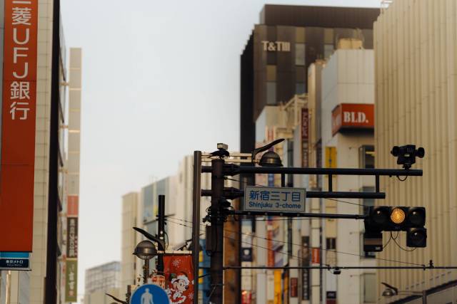 Compressed Shinjuku facades — a red Mitsubishi UFJ sign, a Shinjuku 3-chōme street sign, and a lit amber traffic signal.