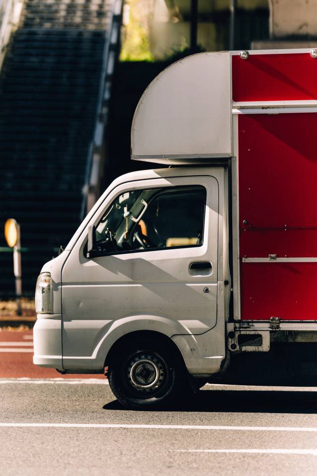 A small white-and-red kei truck parked at the foot of a stairway in hard sunlight.