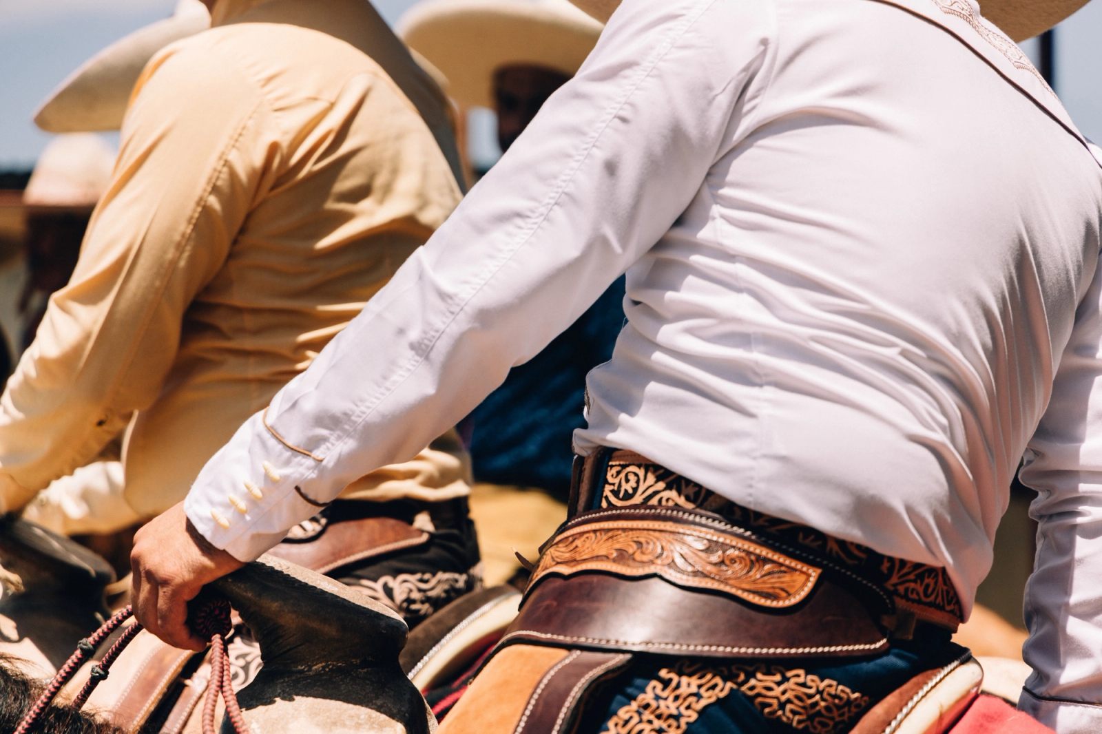 Two charros on horseback from behind, tooled leather saddles and pale hats in hard sun.
