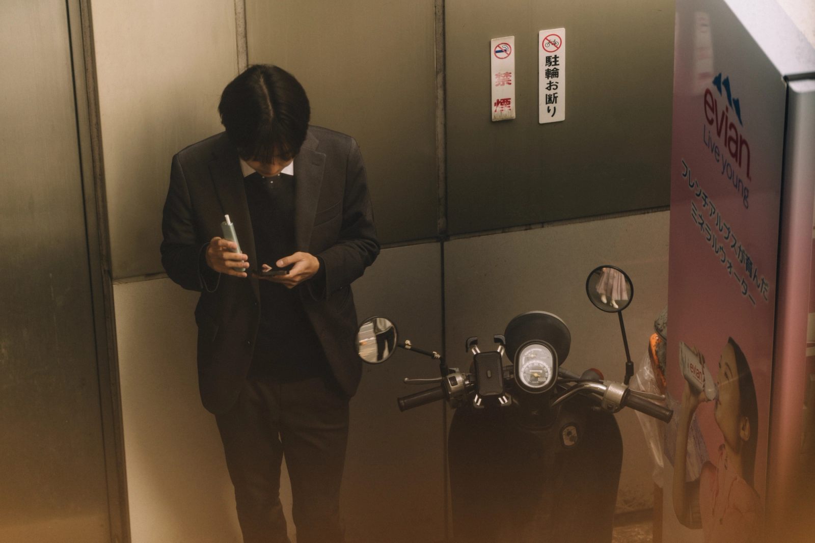 A man in a suit checks his phone in a narrow alcove beside a parked moped and an Evian ad.