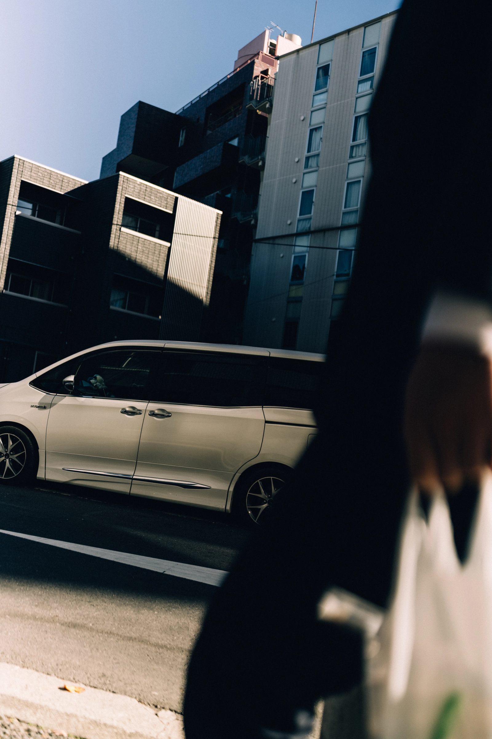 A silver minivan across a sunlit street as a figure with a shopping bag passes in shadow.