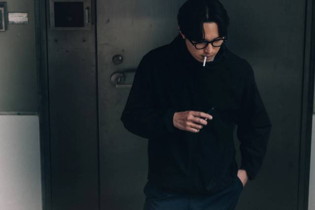 A young man in black lighting a cigarette in front of a steel industrial door.