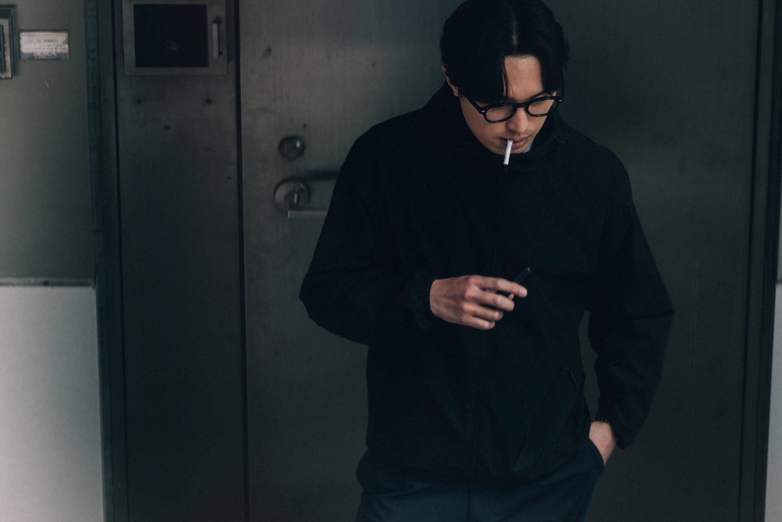 A young man in black lighting a cigarette in front of a steel industrial door.