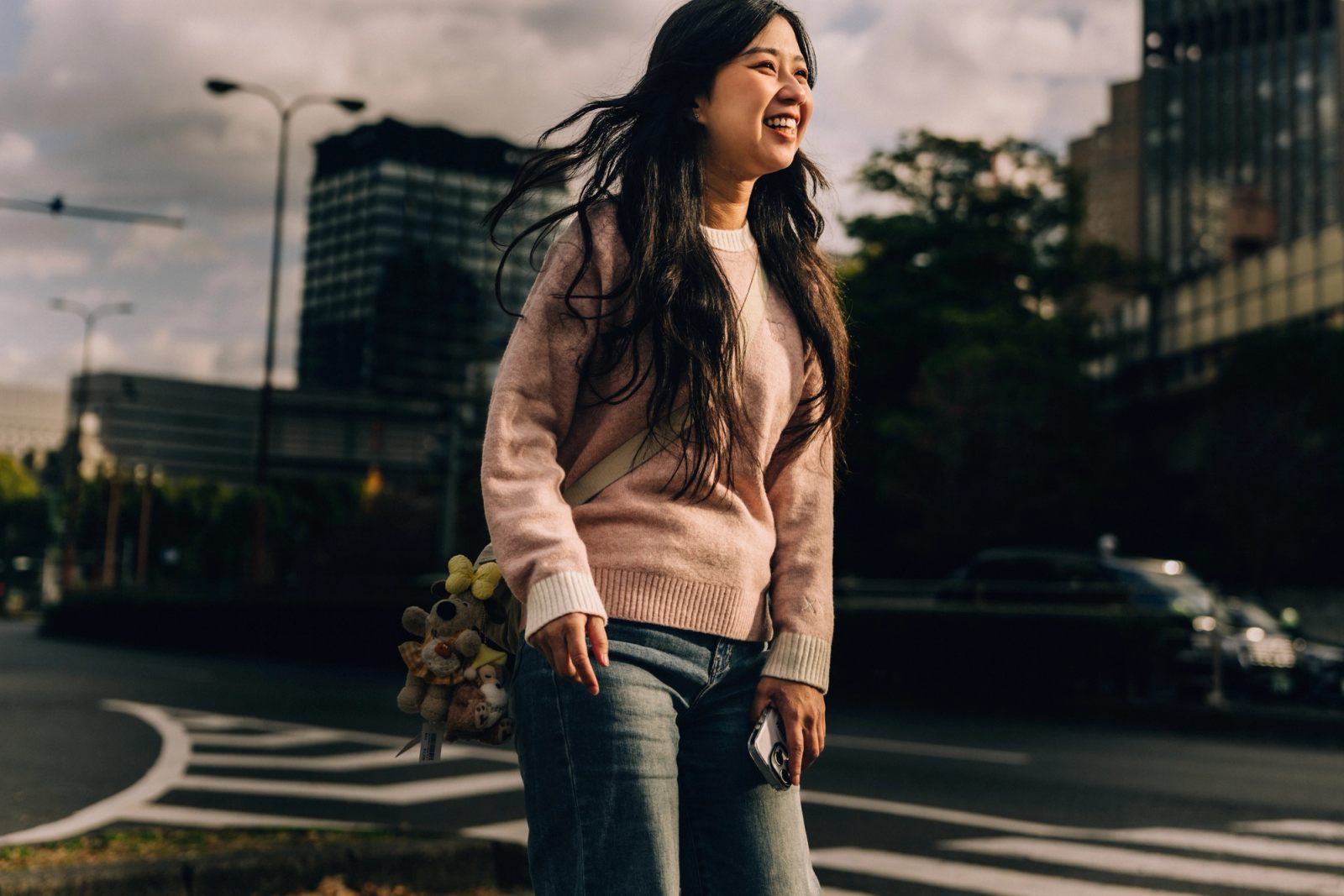 A woman laughs mid-stride on a city street, holding a phone and a bundle of small plush toys.
