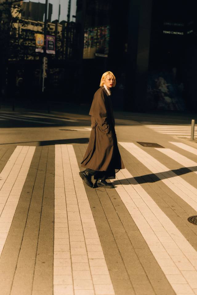 A person in a long coat crossing a wide, sunlit plaza in raking light, cinemas behind.