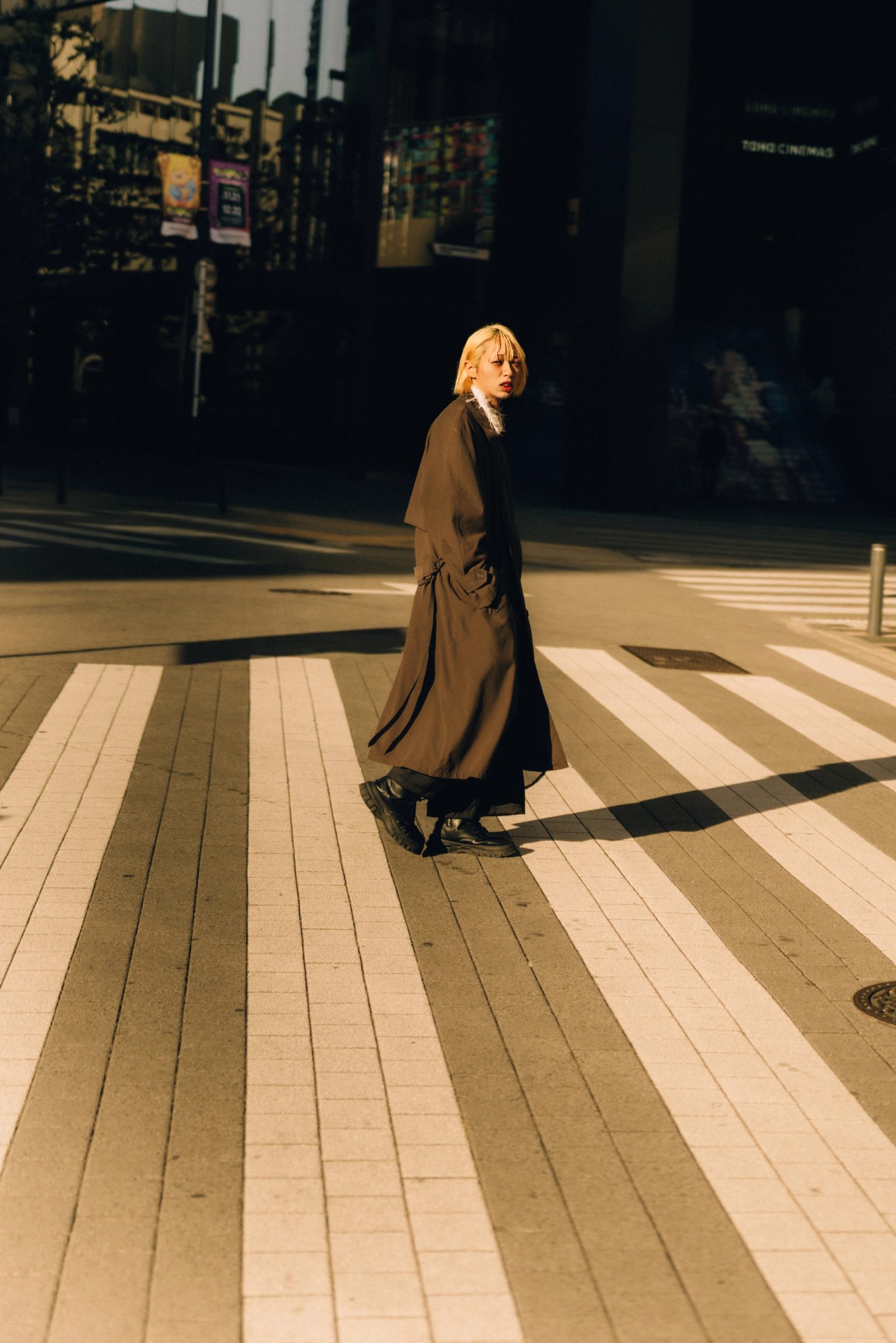 A person in a long coat crossing a wide, sunlit plaza in raking light, cinemas behind.