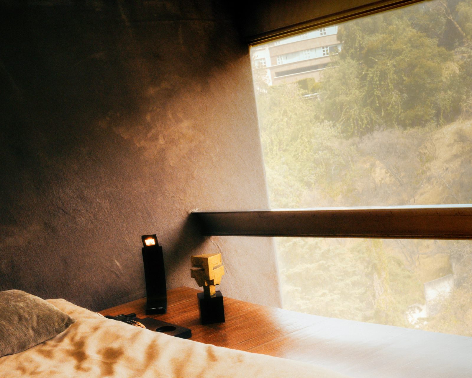 Morning haze over the bed at the angled corner window, a small brass head on the ledge, the ravine green beyond.