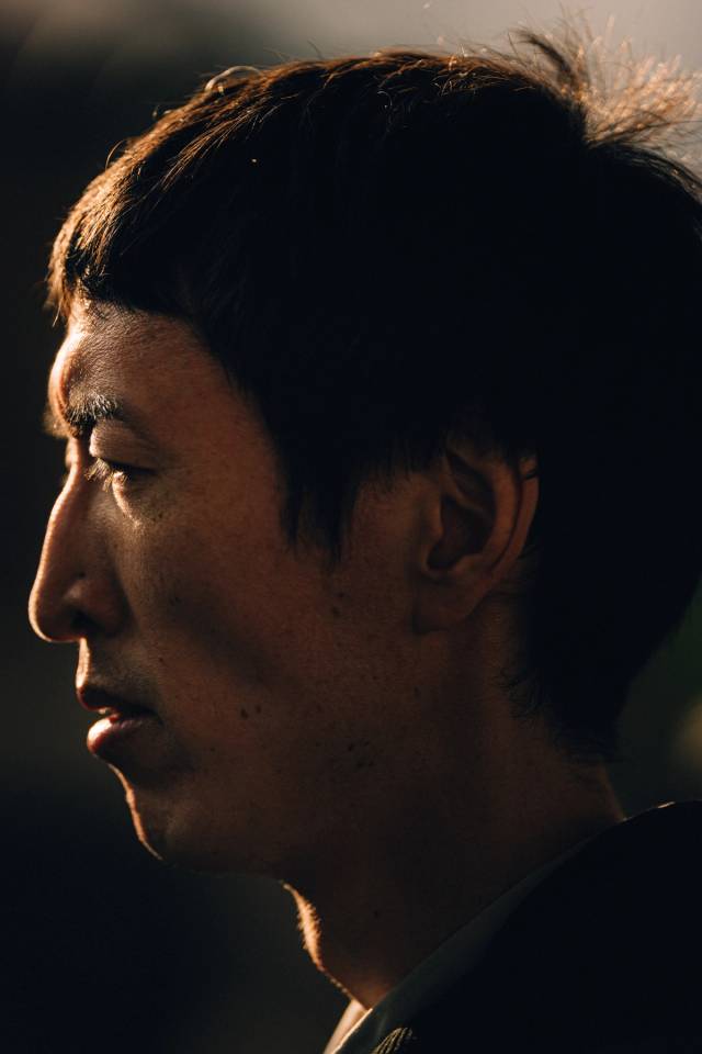 A man in profile, his face lit by a sliver of warm light against darkness.