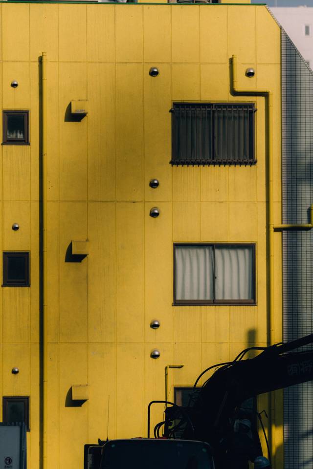 A bright yellow building facade in raking light, dotted with round fixtures, an excavator arm entering the frame.