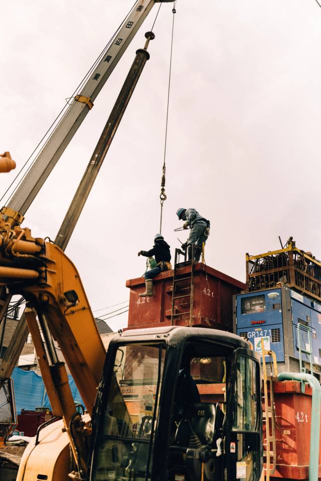Two workers atop a red crane counterweight marked 4.2 t at a construction site under a grey sky.