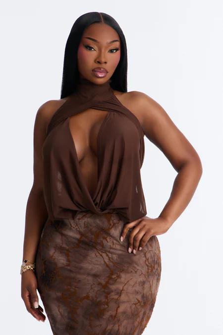 Mujer con top halter-neck café transparente con abertura frontal y falda estampada a juego. Posa sobre fondo blanco.