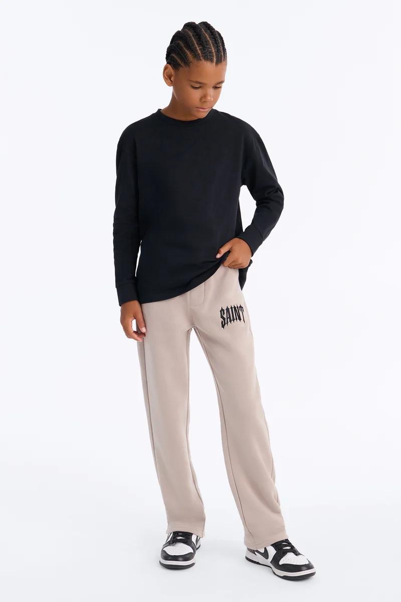 Young boy wearing a black long-sleeve shirt and beige jogger sweatpants with SAINT graphic.
