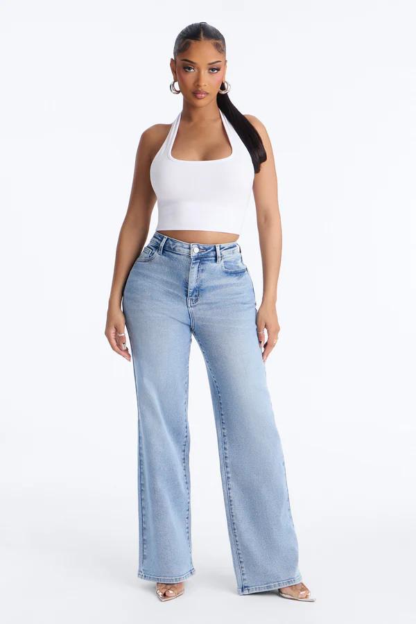 Woman modeling a white halter neck crop top and light wash wide-leg jeans. She has a high ponytail, hoop earrings, and clear heels.