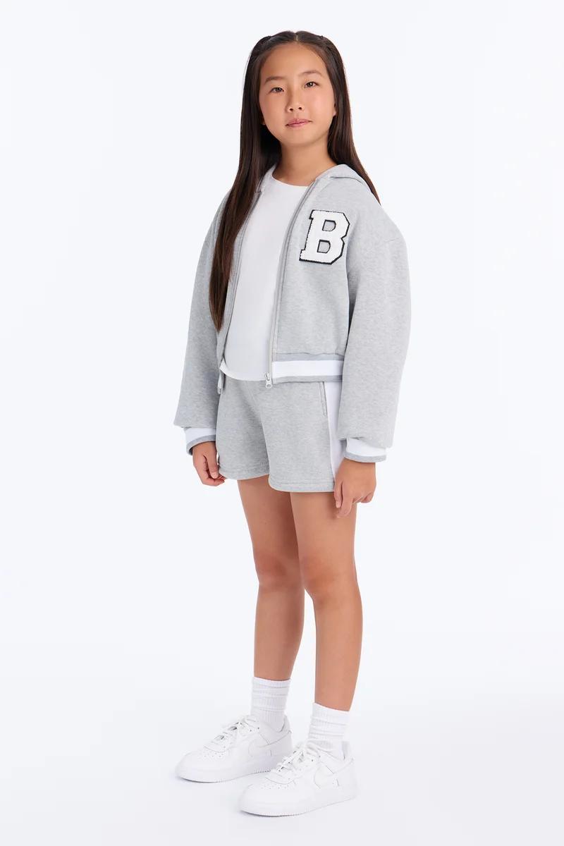 Girls grey varsity zip-up hoodie and matching shorts set with white sneakers.