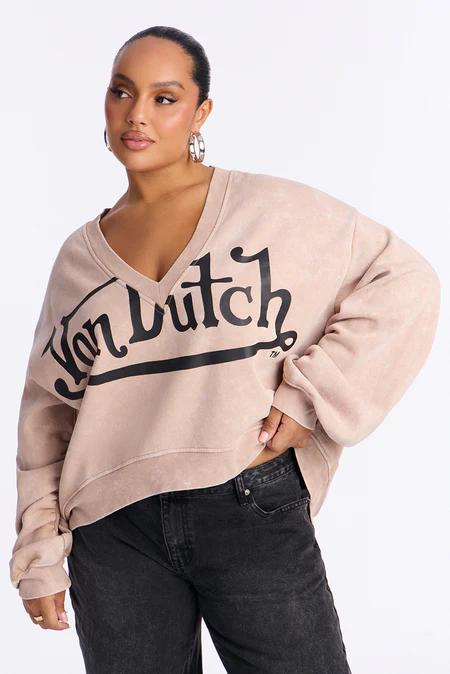 Mujer modelando sudadera beige claro V-neck con logo "Von Dutch" negro y jeans oscuros. Cabello recogido y arracadas plateadas.