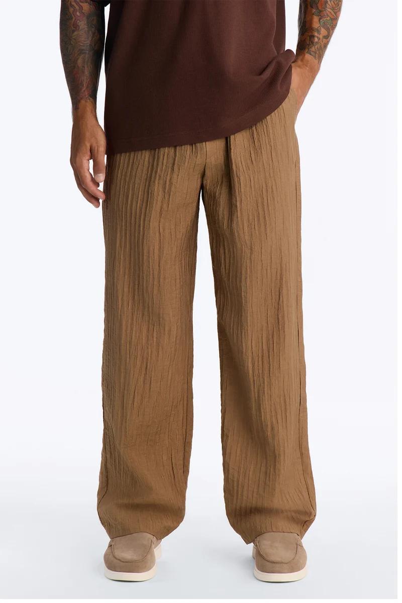Man models relaxed-fit, crinkled brown men's pants and a dark brown top with tan loafers, emphasizing comfortable casual style.