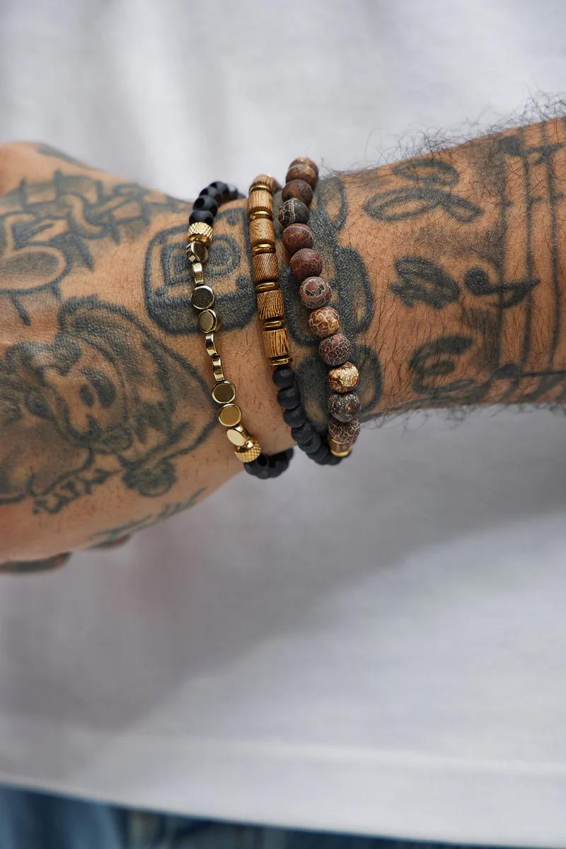 Pulseras de hombre apiladas: doradas, piedra volcánica oscura y piedra natural marrón en un brazo tatuado. Accesorios masculinos.
