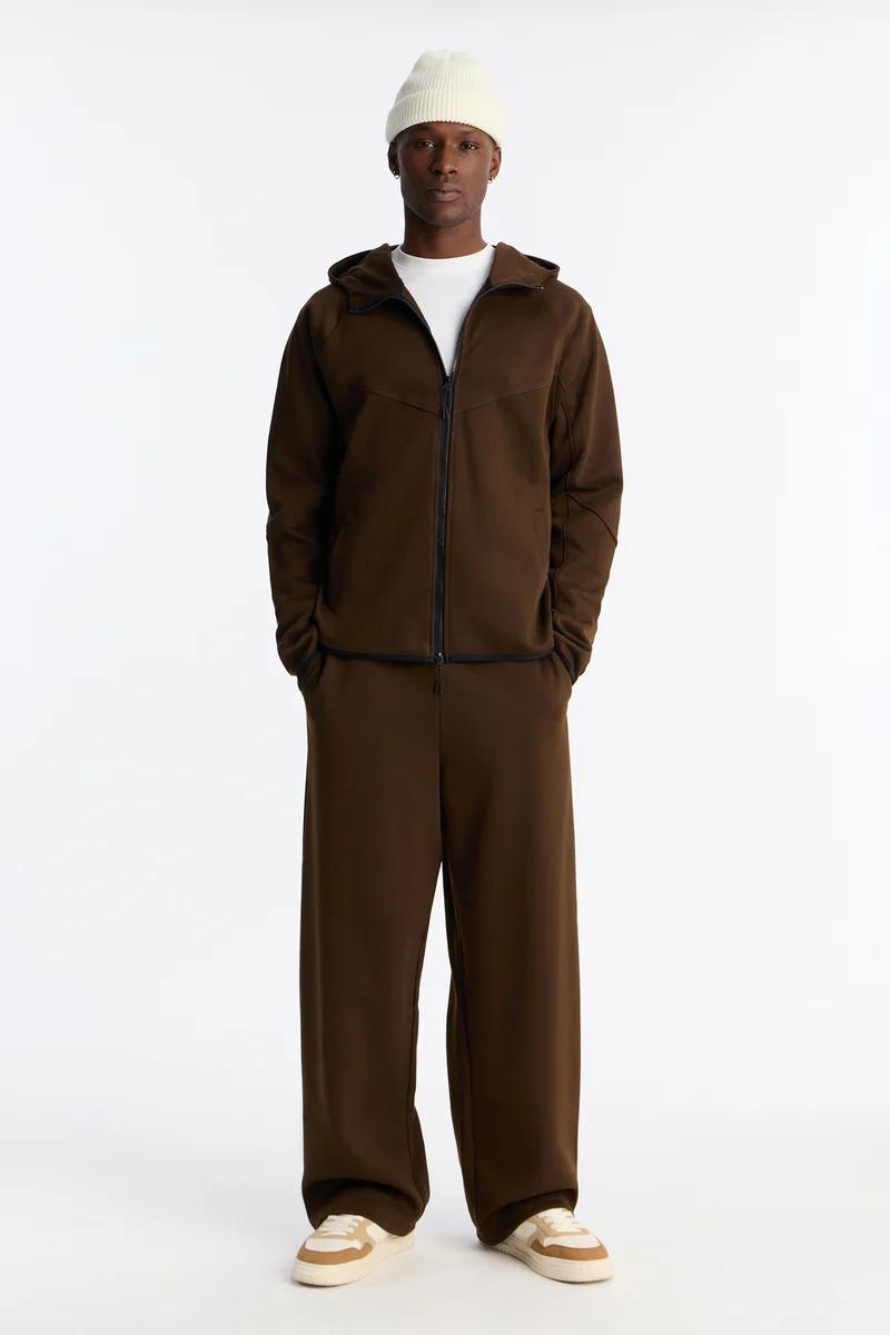 Man wears brown zip hoodie, wide-leg sweatpants, white tee, beanie, beige sneakers. New comfortable men's loungewear collection.