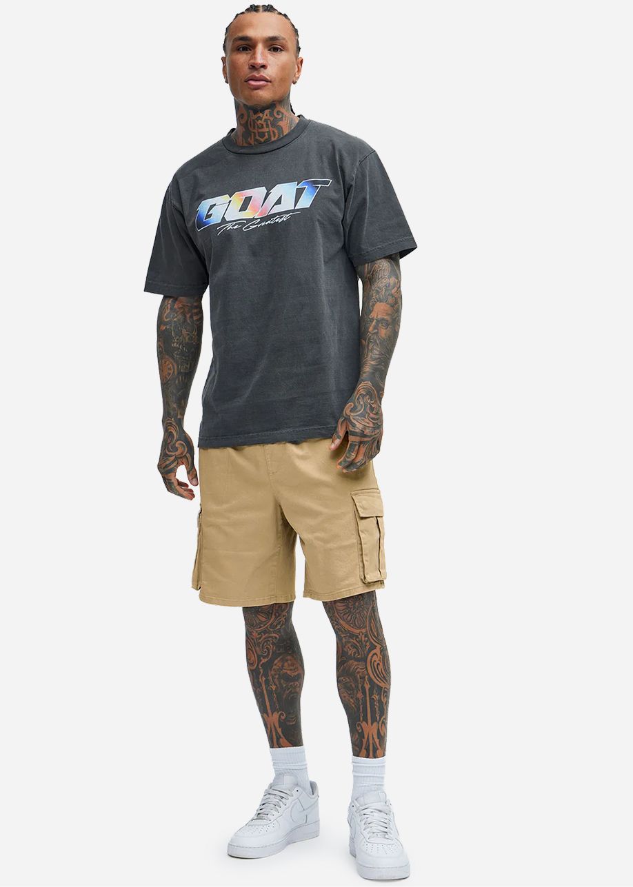 Hombre con camiseta gráfica 'GOAT' gris, bermudas cargo caqui y zapatillas blancas, luciendo moda de verano masculina actual.